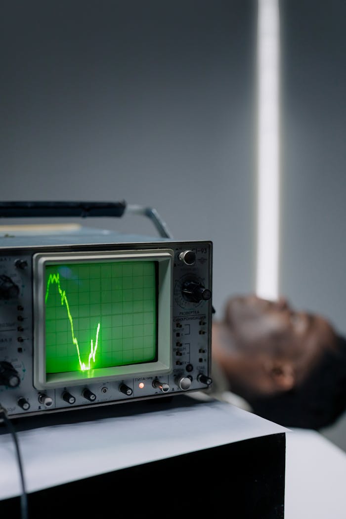 services-01 Close-up of an oscilloscope displaying a pulse signal beside a resting man indoors.